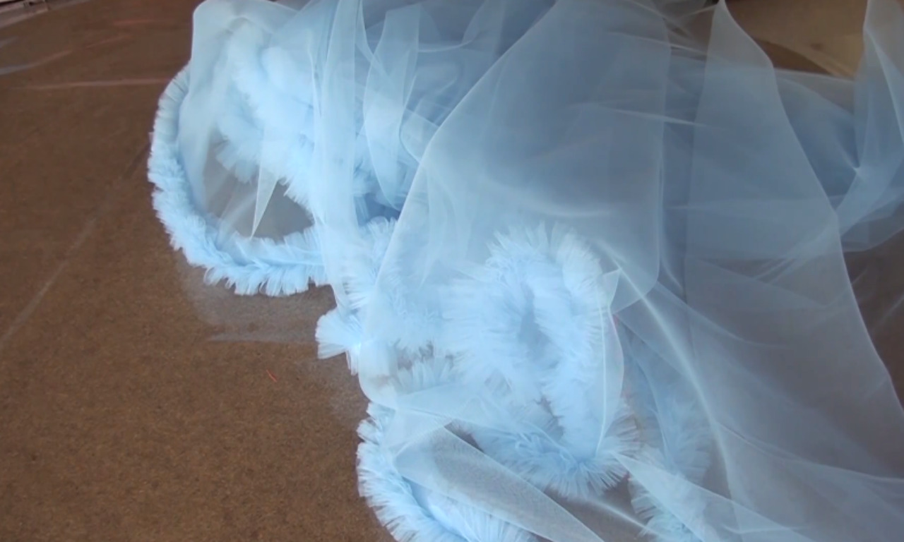 A light blue, sheer fabric, possibly tulle or organza, is draped across a brown surface. The fabric features sections of soft, voluminous ruffles or fringe, creating a wavy, cloud-like texture that contrasts with its smoother, flowing areas.