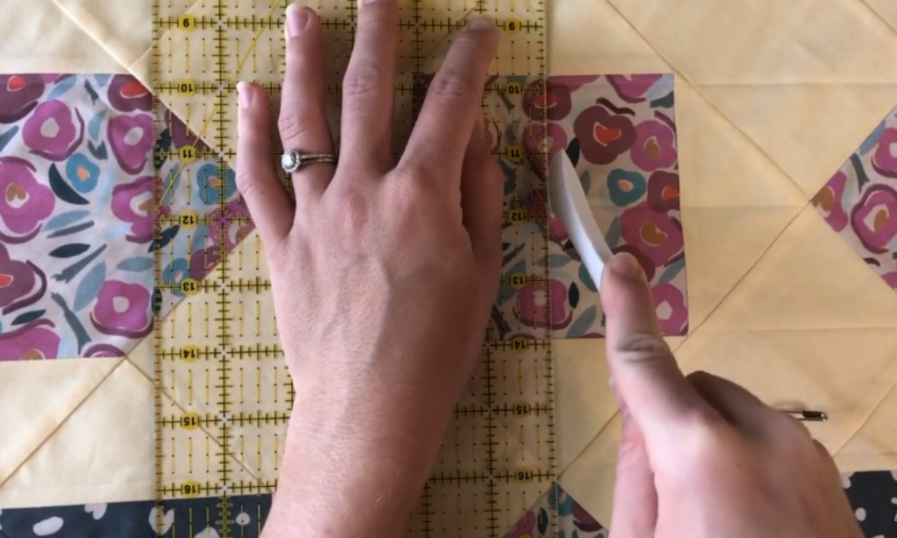hands using a ruler and white marking tool on a quilt block with yellow and floral patterned fabric.