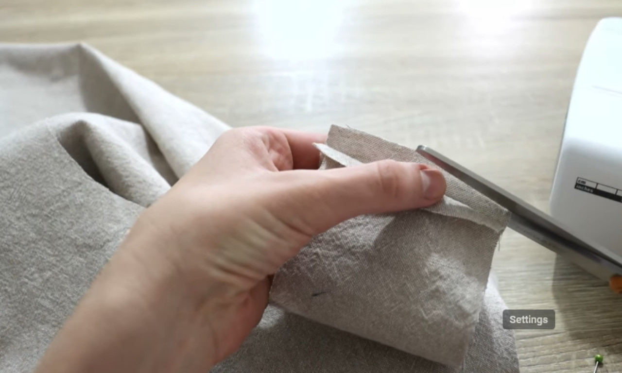a hand holding a folded hem on oatmeal-colored linen fabric and using scissors to snip off the corner tip. A sewing machine sits on the wooden table in the background.