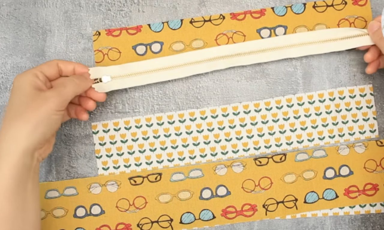two hands holding an open, cream-colored zipper with a silver pull. Below the zipper, on a grey textured surface, lie three rectangular pieces of fabric. The top and bottom pieces are wider, featuring a mustard-yellow background with a repeating print of colorful eyeglasses. A narrower middle strip of fabric has a cream background with a pattern of small yellow tulips.