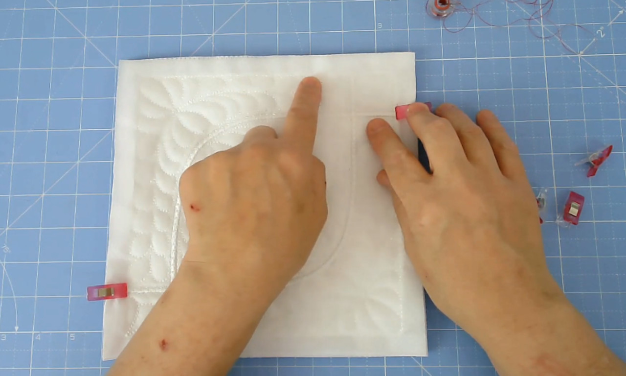 hands working on a white, quilted fabric block on a blue gridded cutting mat.