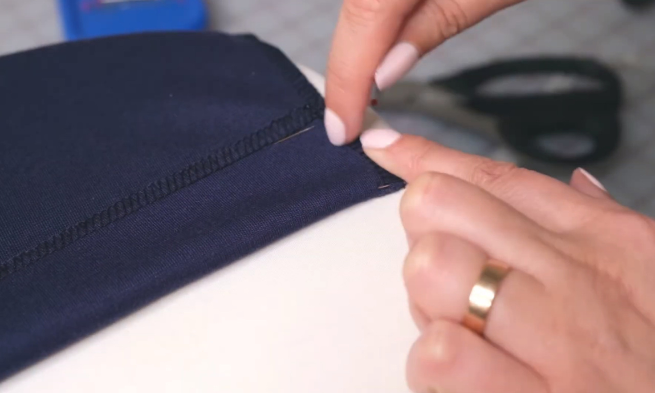A pair of hands carefully trimming the excess thread from a seam on a piece of dark blue fabric using a small pair of scissors. The fabric is laid flat on a white surface, and the person is wearing a gold ring on their ring finger.