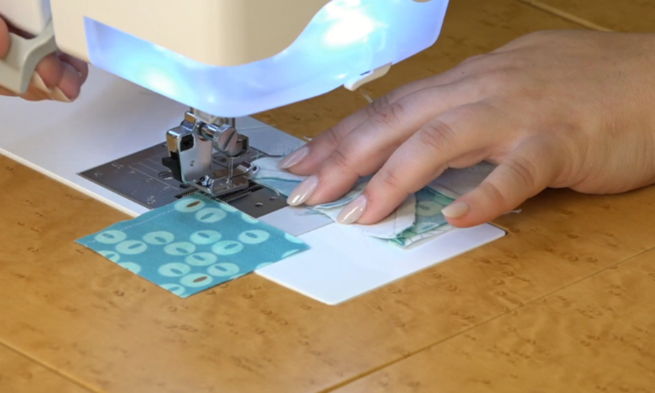 A person is guiding fabric under a sewing machine. The fabric appears to be a blue piece with white circular patterns and a white piece. The sewing machine's needle is positioned over the fabric, and the person's hands are carefully holding the fabric in place on the sewing machine's flatbed.