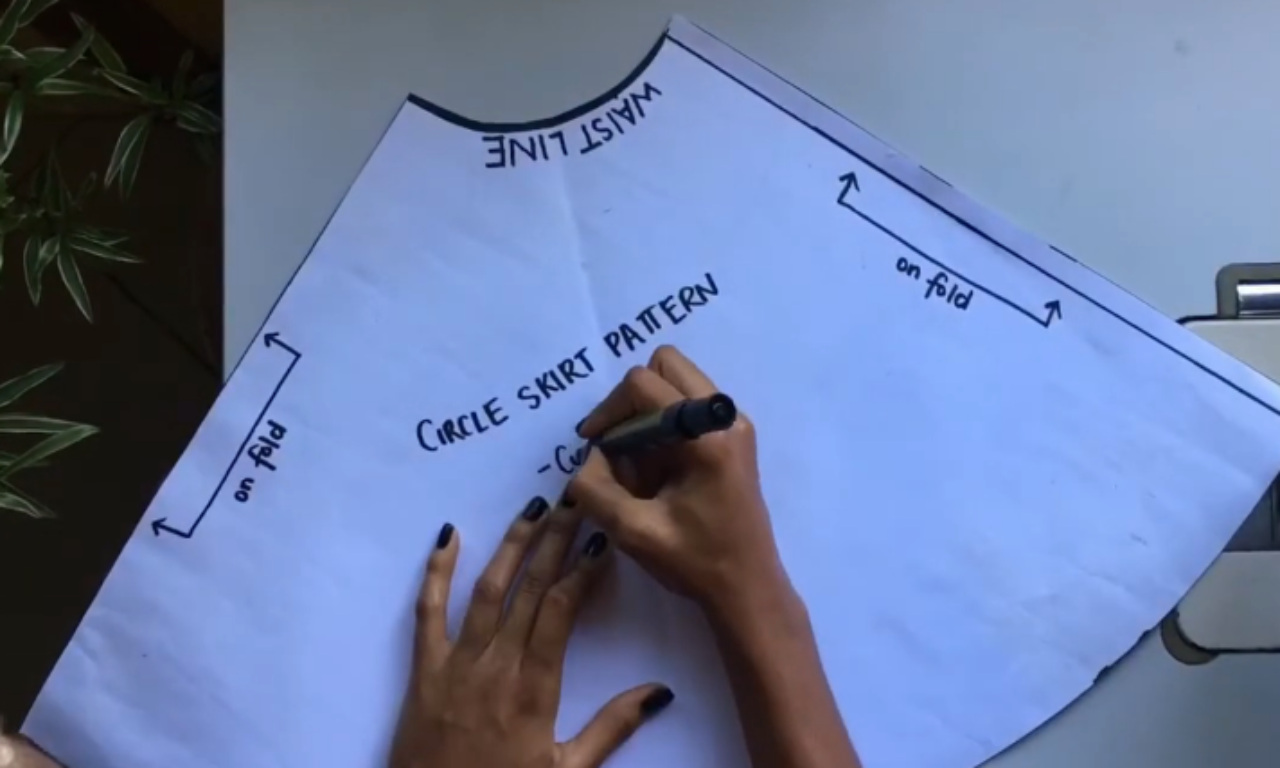An overhead view of a person's hands writing with a black marker on a white paper sewing pattern for a circle skirt. The quarter-circle pattern is labeled "WAIST LINE" on the curved edge and "on fold" on the two straight edges. In the center, the words "CIRCLE SKIRT PATTERN" are written.