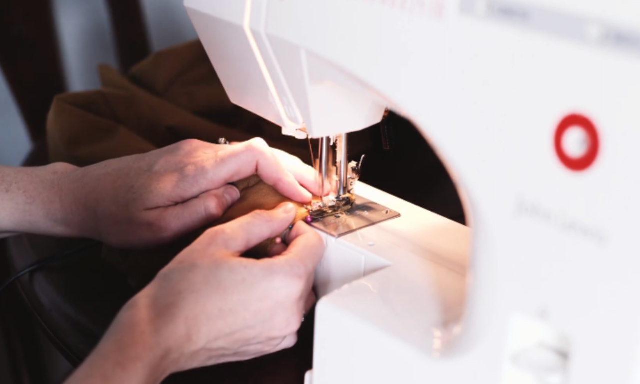a person’s hands guiding brown fabric under the needle of a white sewing machine as they stitch a seam.
