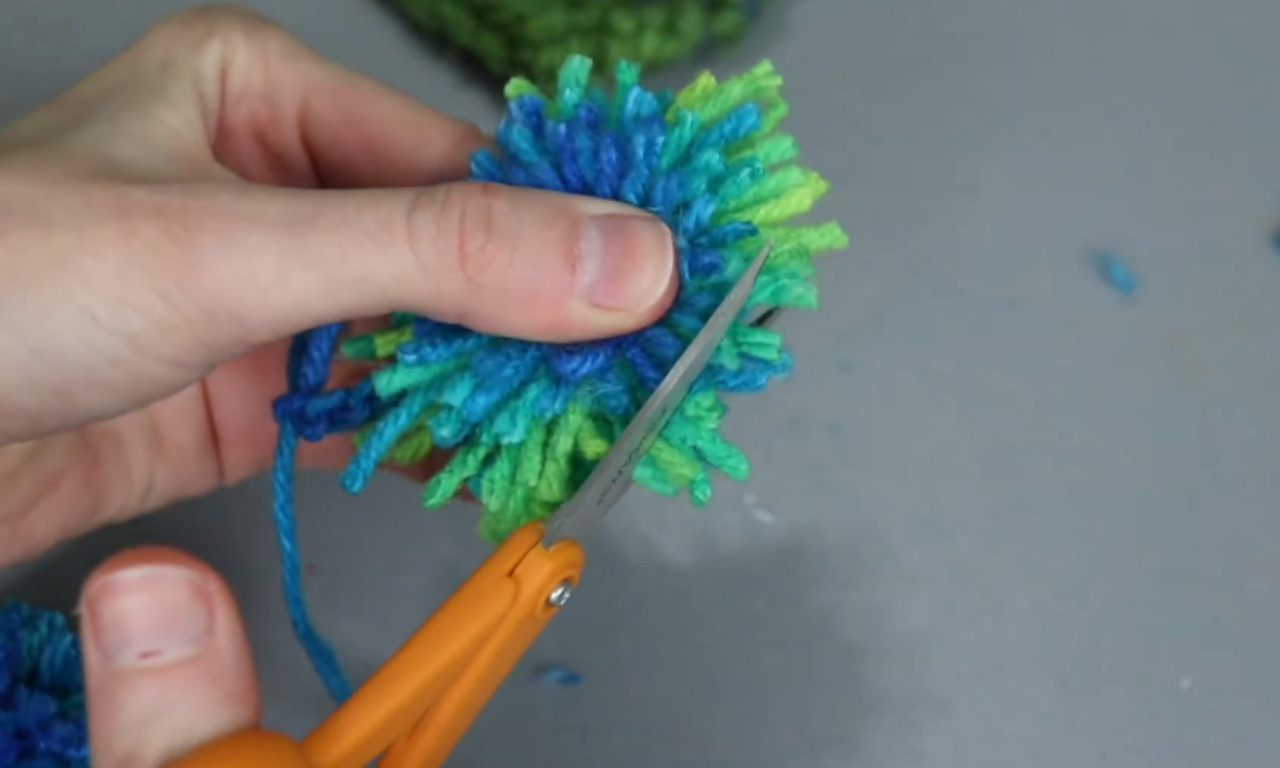 a hand holding a colorful yarn pom-pom. The pom-pom is made of blue, green, and turquoise yarn. The hand is using a pair of orange-handled scissors to trim the yarn, shaping the pom-pom. The background includes a surface with some scattered yarn pieces and a partially visible crocheted or knitted item in similar colors.