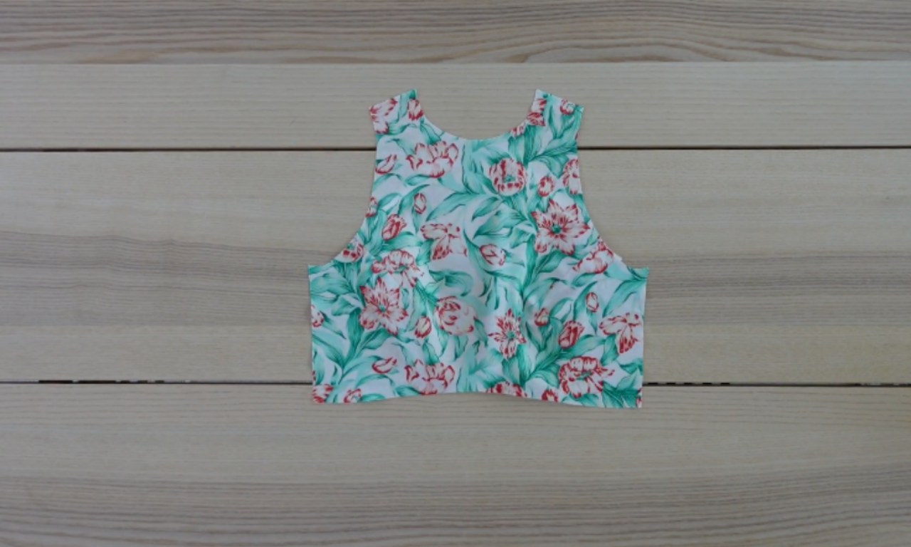A flat piece of white fabric, cut in the shape of a sleeveless crop top, with a repeating pattern of large green leaves and red-orange flowers. It is laid on a light brown wooden surface with horizontal planks.