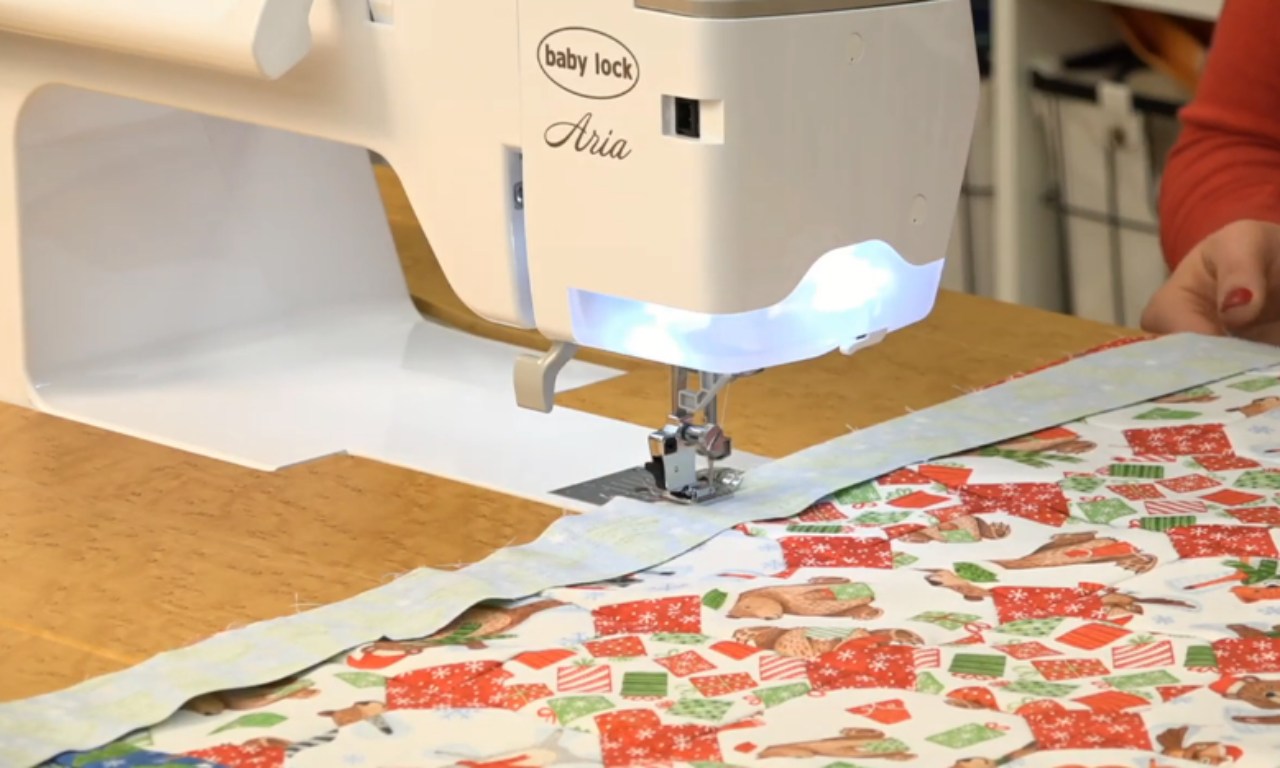 A Baby Lock Aria sewing machine in operation on a wooden table. The machine is white with illuminated stitching area and is currently sewing a colorful Christmas-themed fabric featuring red and green presents, stockings, and gingerbread men. A person's hand is visible at the edge of the frame, guiding the fabric through the machine.