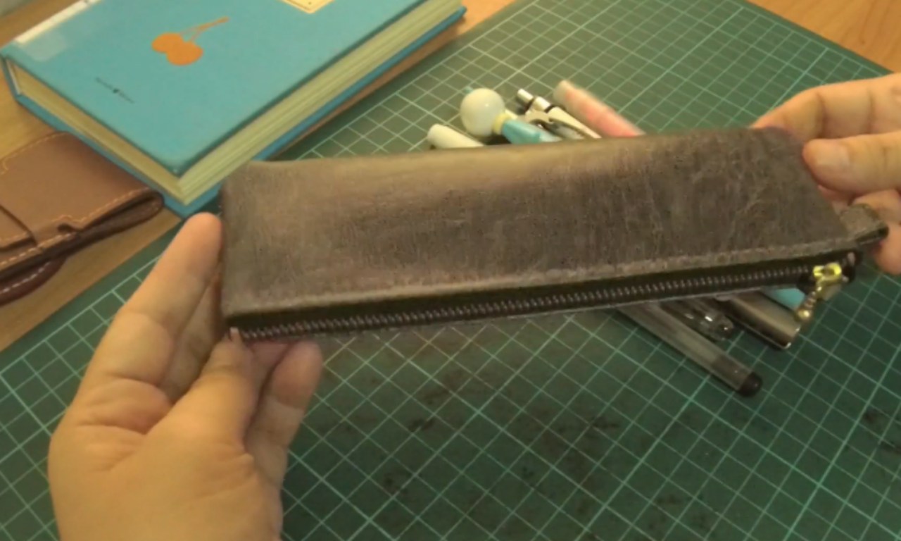 Hands holding a dark leather zippered pencil case filled with pens over a green cutting mat, with a blue book and brown wallet in the background.