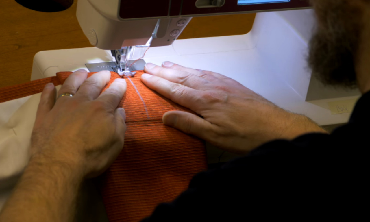 a person using a sewing machine. The individual is guiding a piece of bright orange fabric under the needle of the sewing machine, carefully holding the fabric with both hands to ensure it is sewn evenly. The sewing machine is well-lit, and the person appears to be focused on their task. The scene captures the detailed process of sewing, highlighting the precision and care involved in the craft.