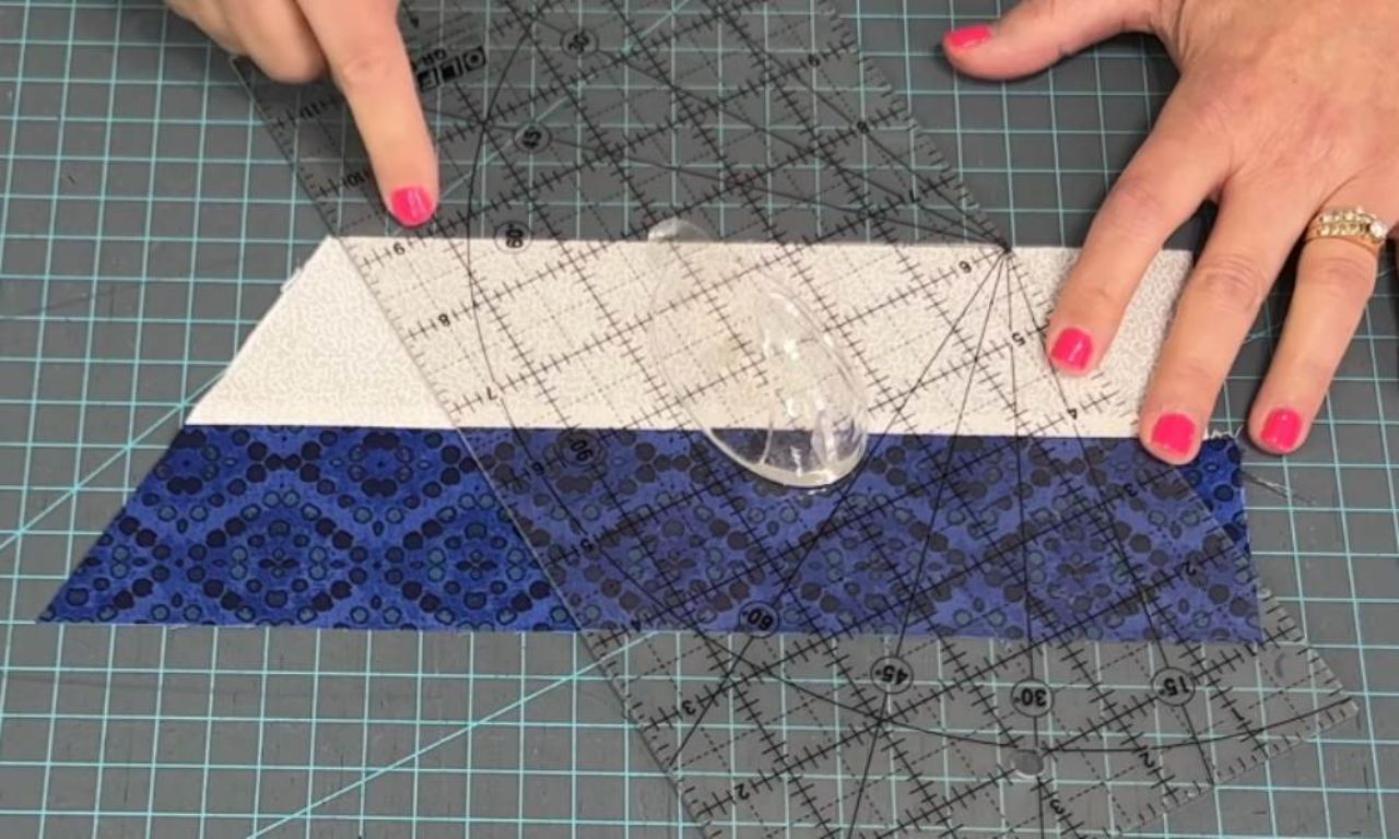 A close-up shows hands with pink nail polish using a clear acrylic quilting ruler with grid lines and angle markings to measure and prepare a two-tone fabric piece on a dark gray grid cutting mat. The fabric has a light gray patterned strip on top and a dark blue patterned strip below. A clear oval ruler grip is centered on the ruler to hold it steady.
