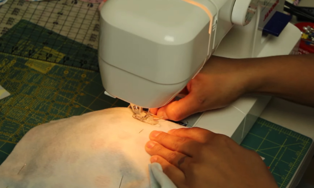 a person using a sewing machine to work on a piece of fabric. The person's hands are guiding the fabric under the sewing machine's presser foot, with the needle visible as it stitches. The sewing machine is placed on a green cutting mat with grid lines, and various sewing tools and materials are visible in the background.