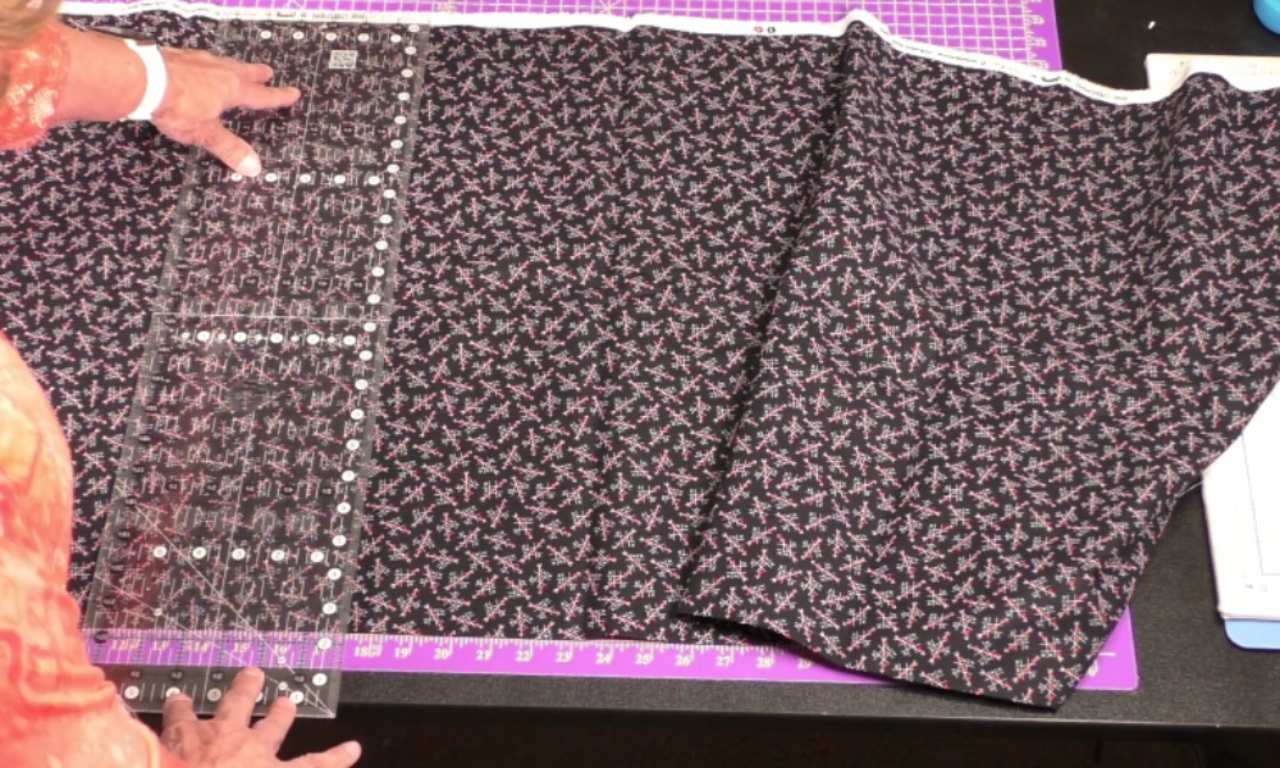 black cotton fabric covered in small pink floral designs. A clear acrylic quilting ruler with grid markings is positioned on top of the fabric, which is laid out on a purple self-healing cutting mat. Hands with bracelets are visible, holding the fabric taut.