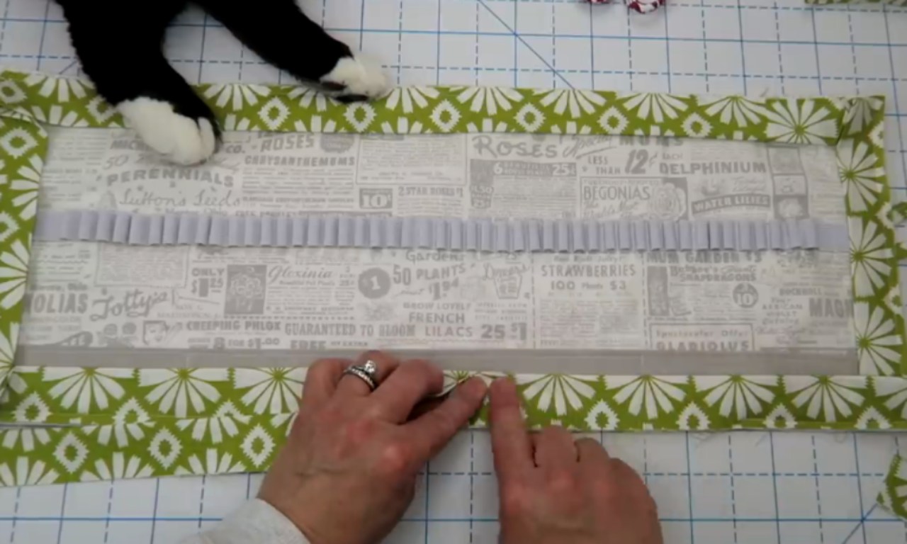 hands tying a ribbon around a rectangular craft project. The project features a background of vintage-style newspaper print, layered with a ruffled lavender paper trim, and a green floral patterned fabric border. A black cat paw is visible in the upper left corner.