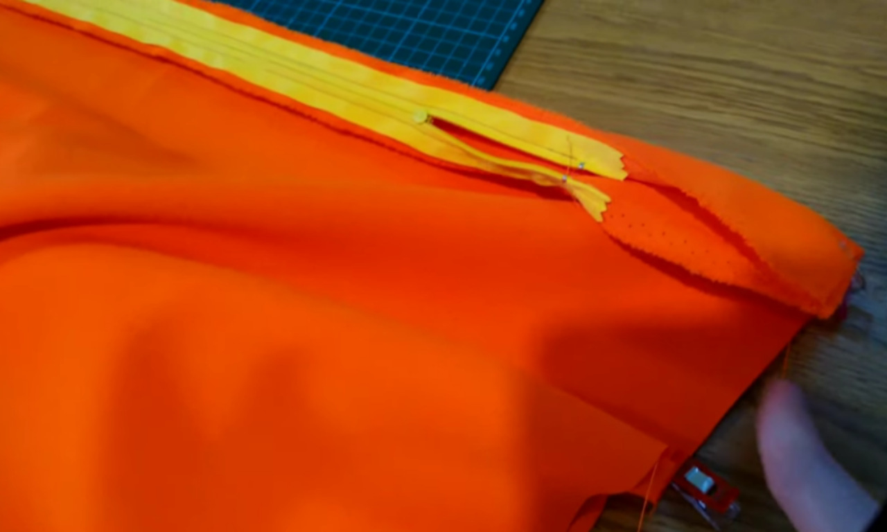 a bright orange fabric panel laid flat on a wooden tabletop, with a yellow zipper pinned along its raw edge using sewing clips; a green cutting mat peeks out at the top, and a hand is visible at the lower right corner, guiding the fabric.