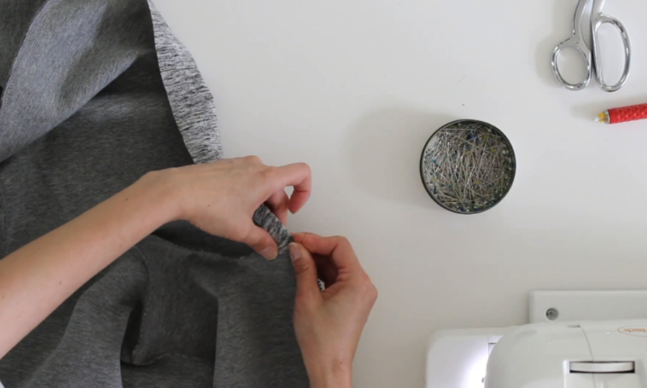 hands pinning the edge of grey fabric on a white work surface next to a sewing machine, a magnetic pin cushion, and scissors.