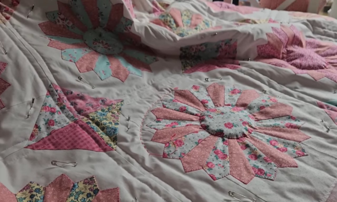 A person's hands and quilting tools (needle, thread, thimble) are visible as they hand-quilt the fabric.