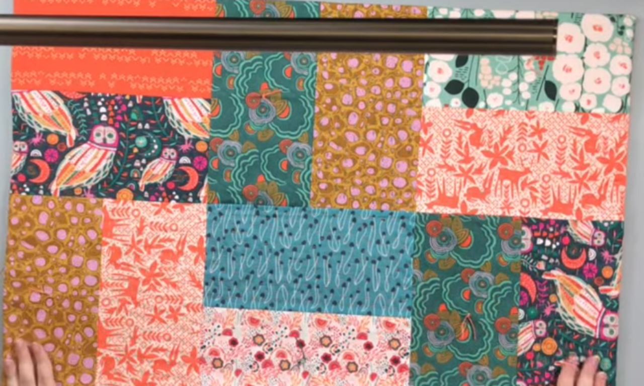 A vibrant patchwork textile composed of various rectangular blocks of fabric. The patterns include colorful owls on a dark blue background, coral deer and foliage, pink and white flowers on a light green background, abstract gold and pink spots, teal wavy lines, and dark green organic swirls. A dark metal bar runs horizontally across the top, and a person's hands are gently holding the bottom corners of the fabric, which is laid out flat against a light background.