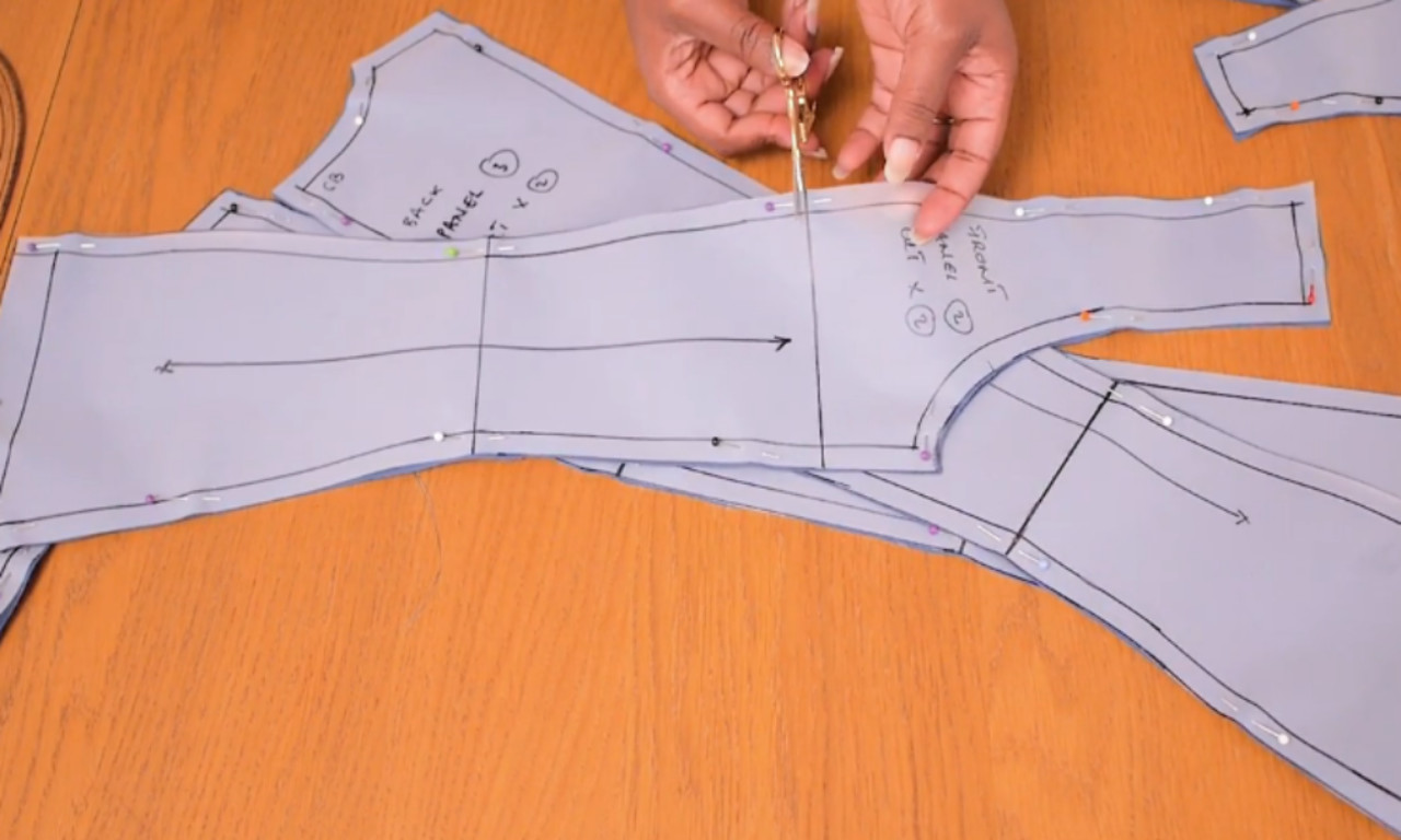 a pair of hands using scissors to cut a light blue paper sewing pattern, detailed with black lines and pins, on a wooden table.