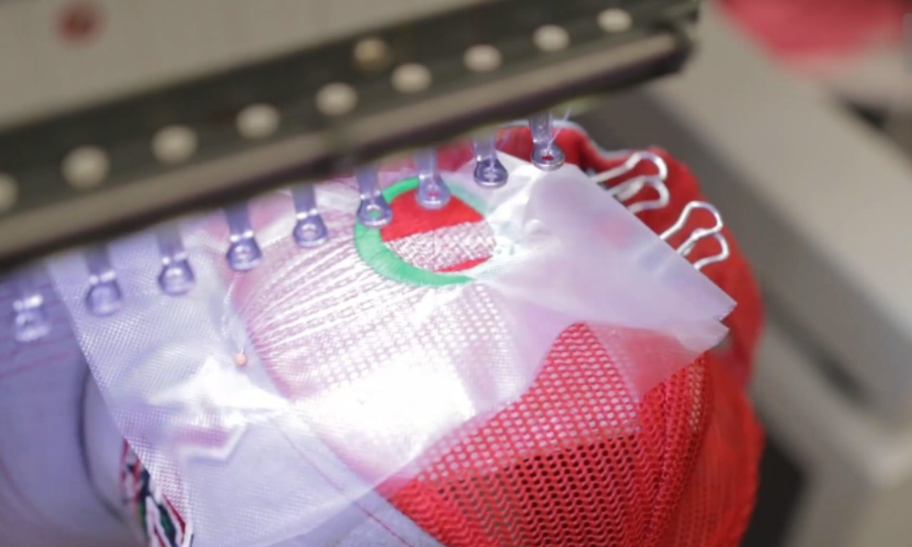 An automated embroidery machine with multiple needles stitches a logo onto the front of a red trucker hat. The hat is held in a frame, and a clear stabilizer sheet covers the active stitching area.