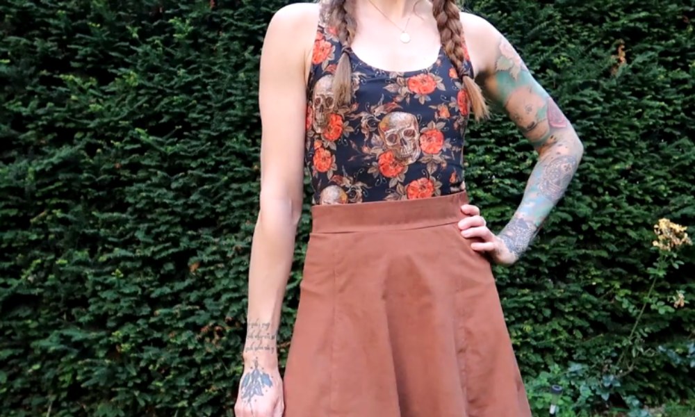Woman standing in front of a green hedge, wearing a black tank top with a skull and floral pattern, a brown skirt, and displaying tattoos on her arms.