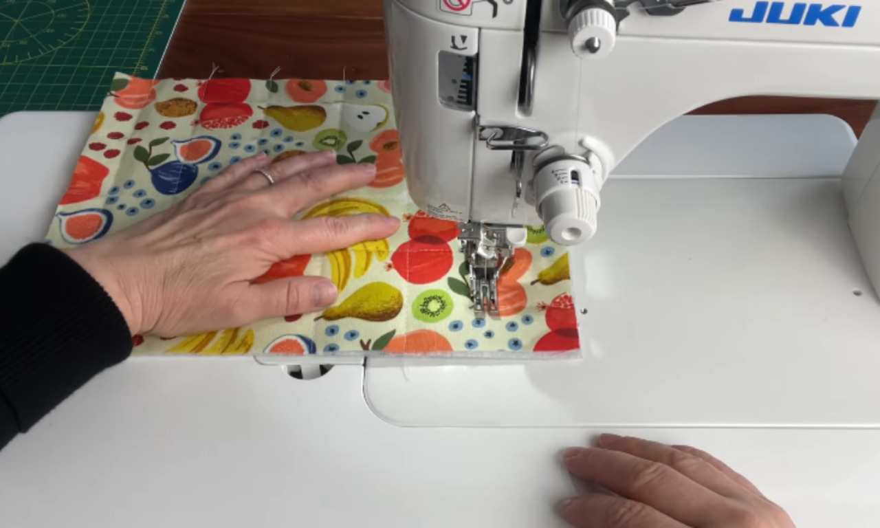 A person's hands working on a colorful piece of fabric with a fruit pattern on a Juki sewing machine, with a green cutting mat and wooden surface visible in the background. The person's left hand guides the fabric while their right hand steadies it, as they sew a design onto the fabric.