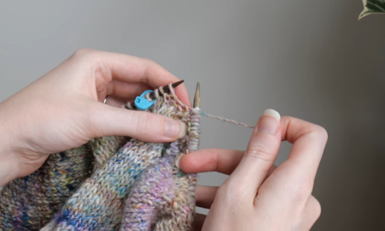 A person is shown working on a multicolored knitting project. Their hands are visible, holding knitting needles and yarn. The person is focused on a specific part of the project, indicated by a blue marker. The background is a plain gray wall, with a hint of a plant visible in the top right corner. The person's fingernails are neatly trimmed and painted white.