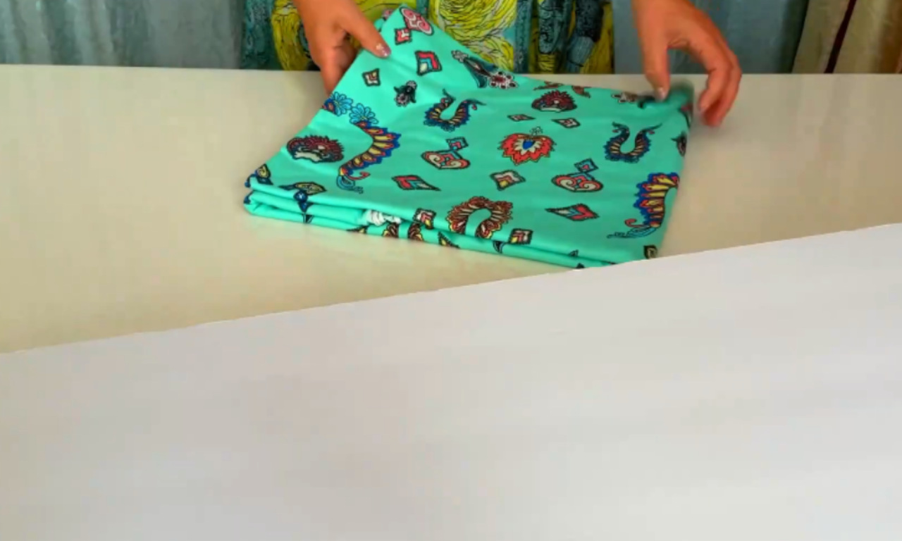 a pair of hands folding a piece of fabric with a colorful, patterned design. The fabric features various playful and vibrant motifs, including cartoon-like characters and symbols. The hands are carefully aligning the edges of the fabric, likely in the process of folding it neatly. The background includes a light-colored surface and a glimpse of a painting or decorative wall hanging.