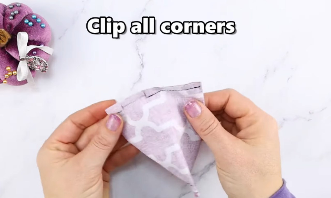 A pair of hands holds up a light purple and white patterned piece of fabric, showing a freshly sewn corner with visible black stitching. The fabric appears to be part of a larger project, possibly inside out, ready for trimming. Text overlaid on the image reads "Clip all corners." In the upper left corner, a purple velvet pincushion adorned with various pins and a white ribbon is visible. The scene is set on a bright white marble surface.