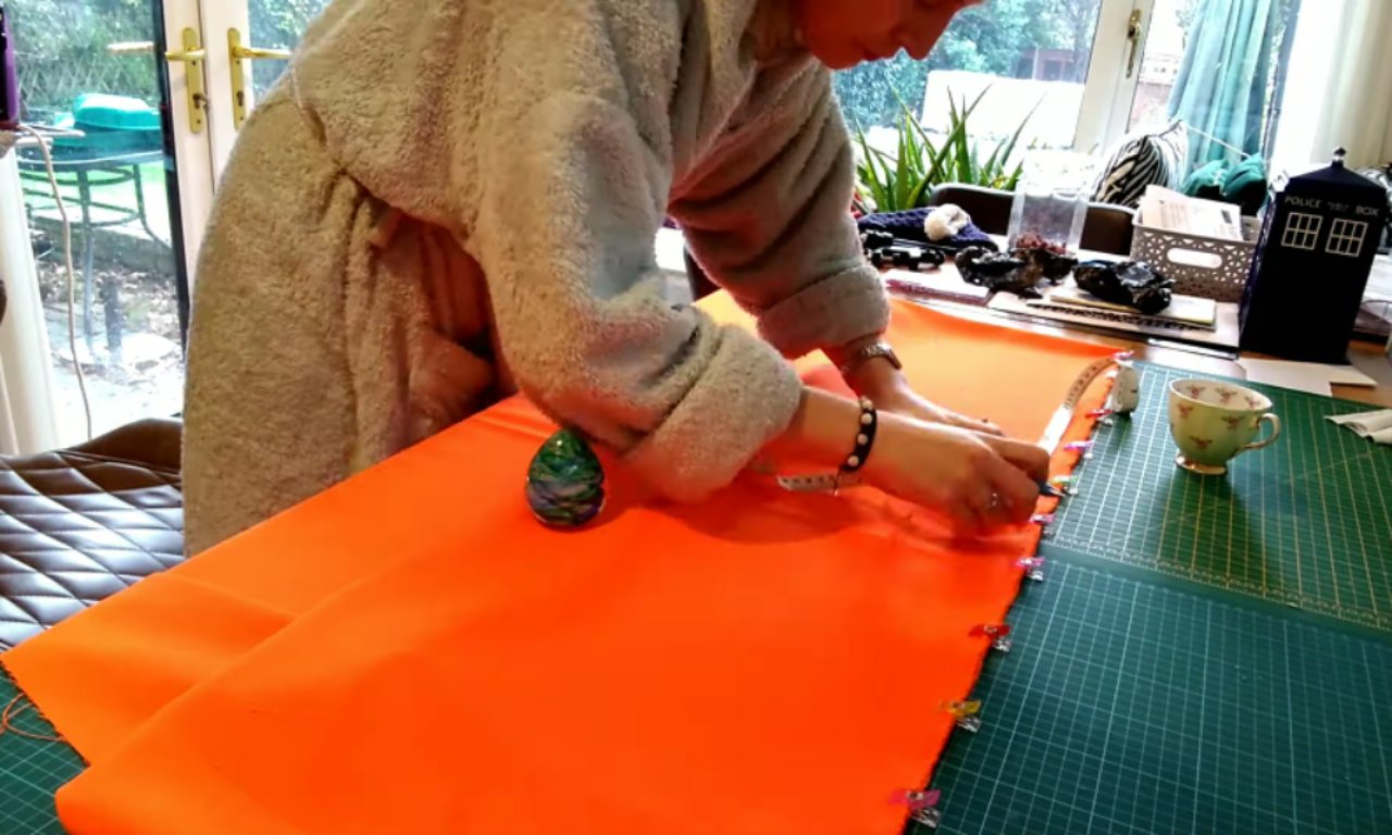 A person wearing a grey fleece is pinning a large piece of bright orange fleece fabric to a green self-healing cutting mat. A decorative dragon egg sits on the fabric, and sewing supplies and a Doctor Who TARDIS figurine are visible in the background.