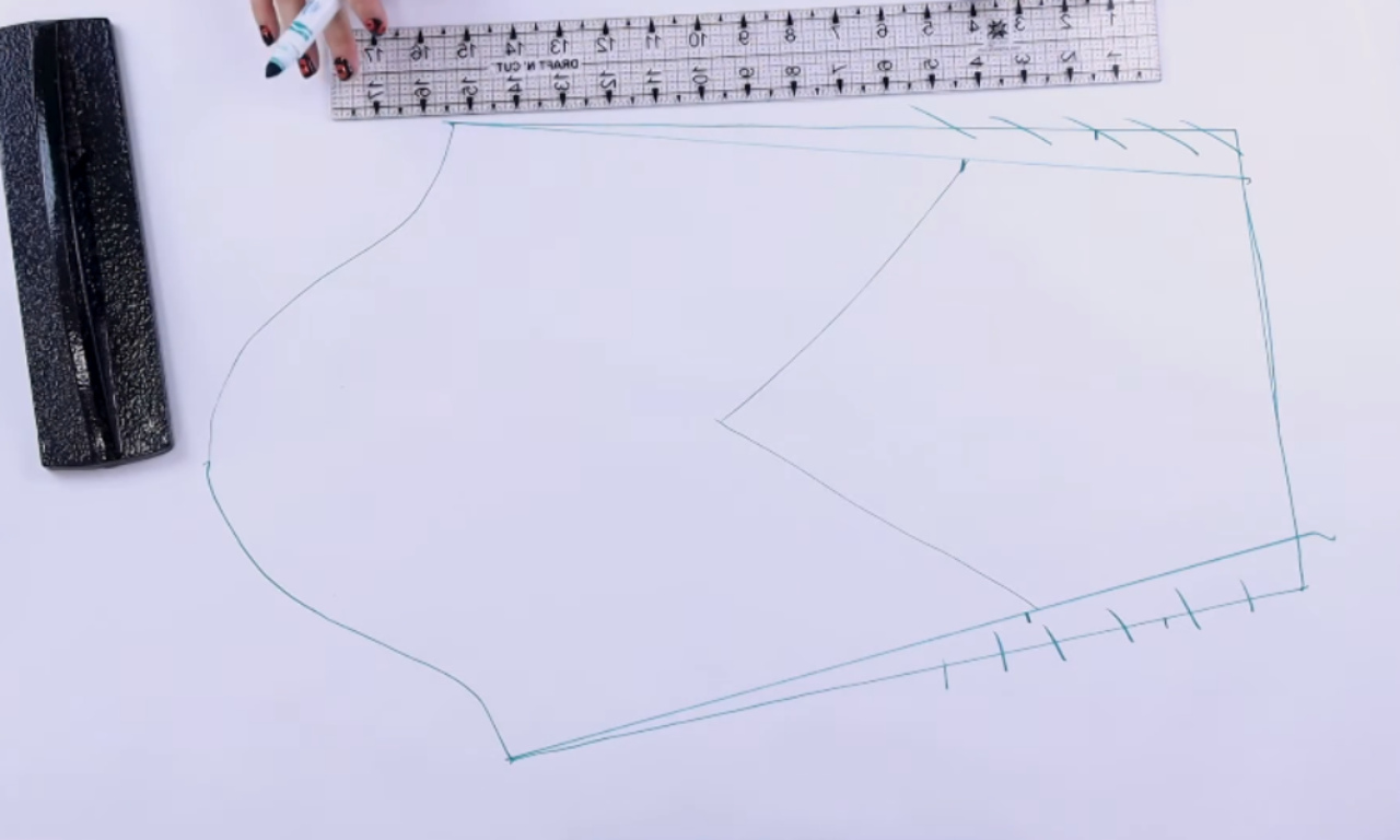 A hand with dark-painted fingernails holds a marker over a ruler and a piece of paper with a partially drawn pattern for sewing or fashion design. The pattern has been drawn in teal and includes markings for cutting and shaping fabric. A black case sits to the left of the paper.