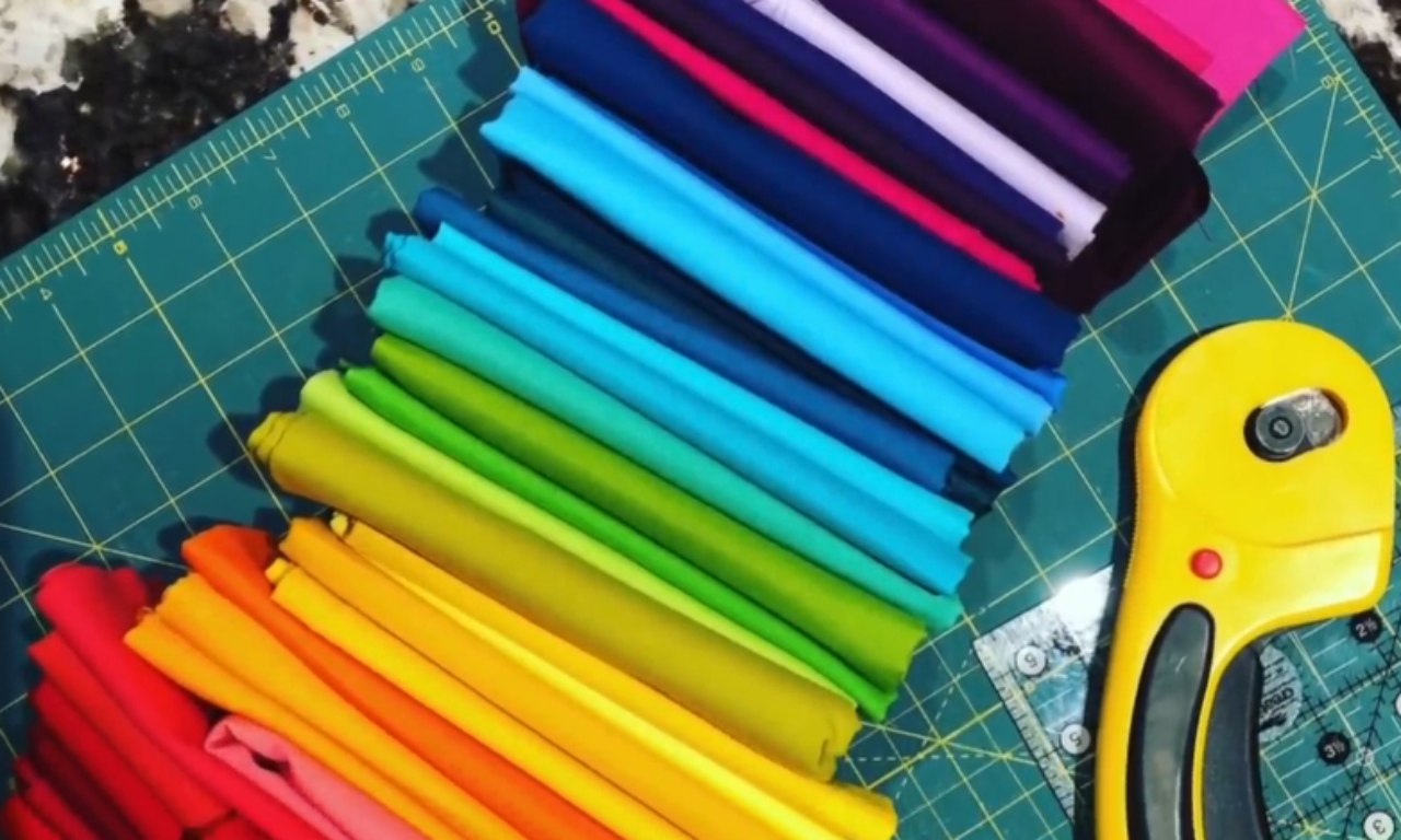Two stacks of folded fabric strips in a rainbow of colors, arranged on a green cutting mat with a yellow rotary cutter and clear ruler.