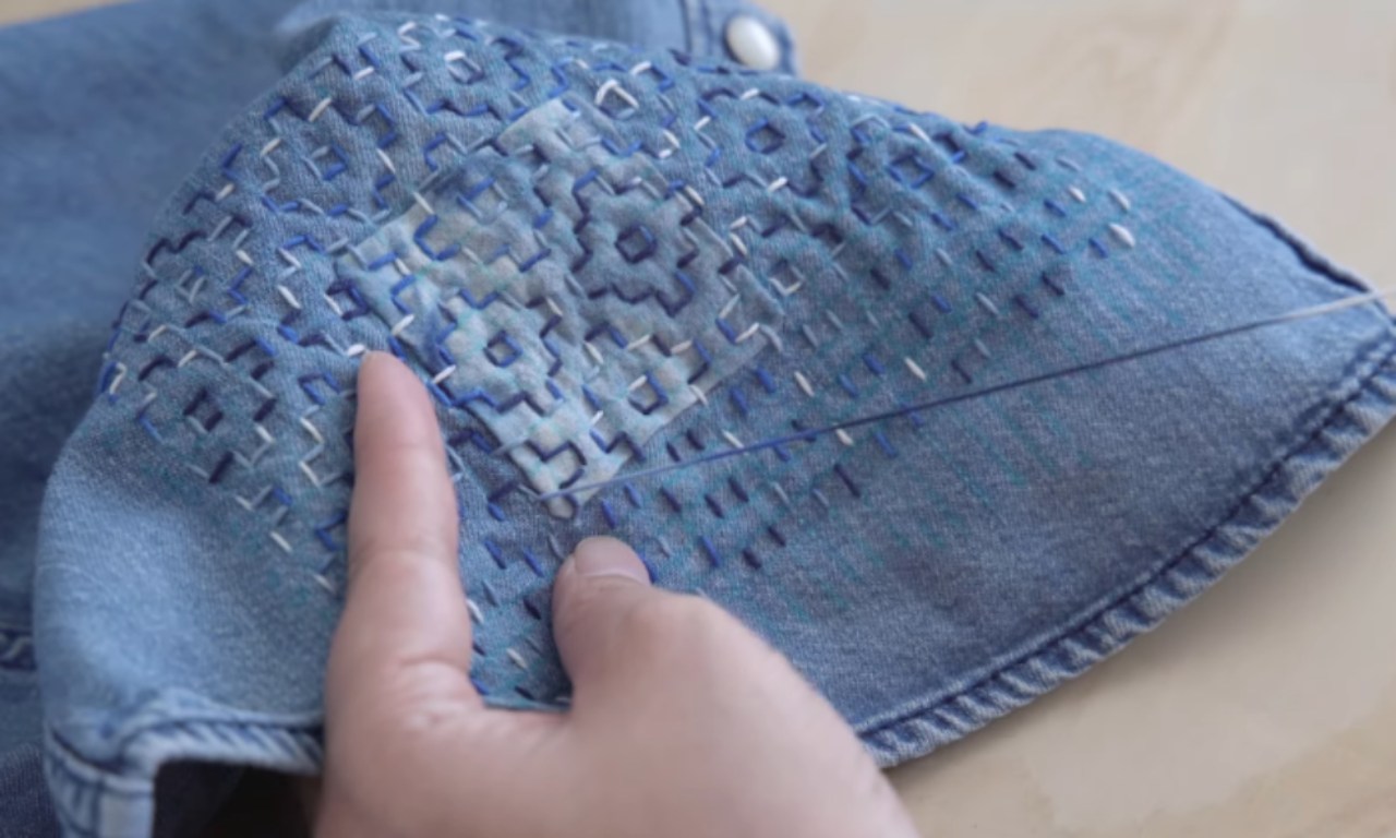 Close-up of a hand holding light blue denim fabric being stitched with dark blue and white thread in a geometric Sashiko pattern.