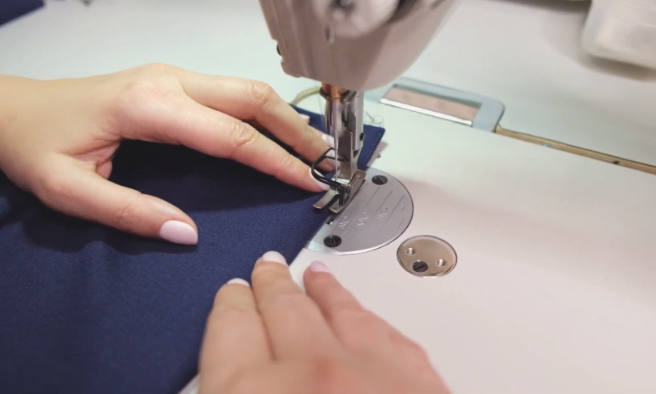 hands guiding dark blue fabric through a sewing machine as it stitches along the edge.