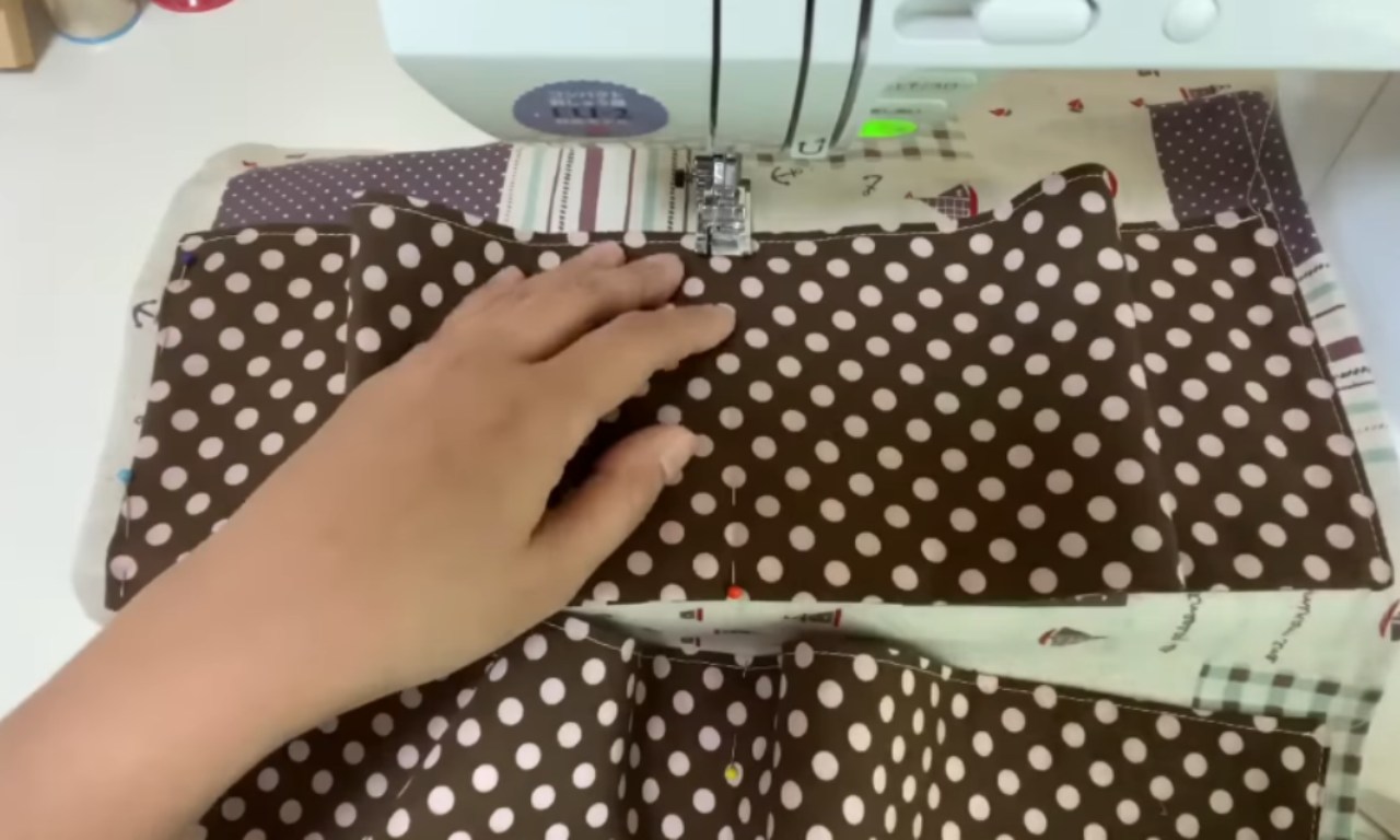 A sewing machine in action with a hand guiding brown polka-dotted fabric through the presser foot. The fabric appears to be part of a sewing project featuring brown fabric with white dots being stitched together with other patterned fabrics visible underneath. The machine's needle is positioned to sew a straight seam across the polka dot material.