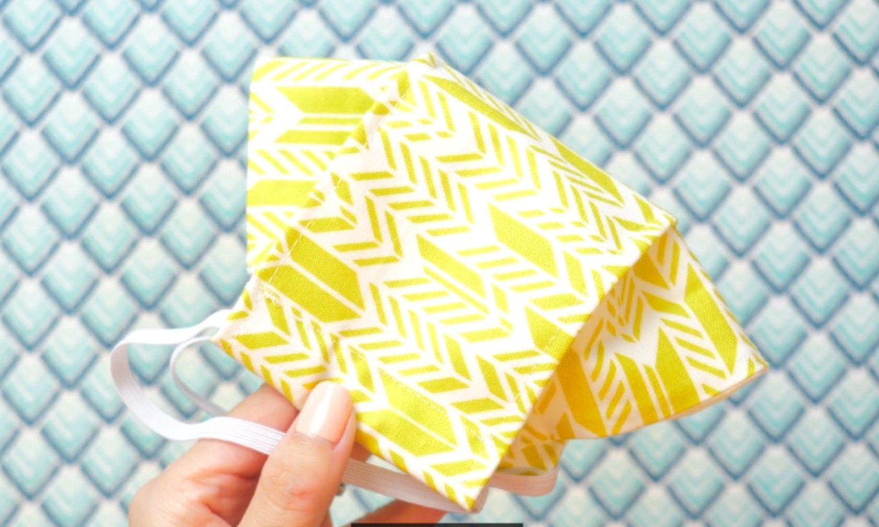 A hand holding a yellow and white patterned face mask with elastic straps against a blue and white diamond-patterned background.