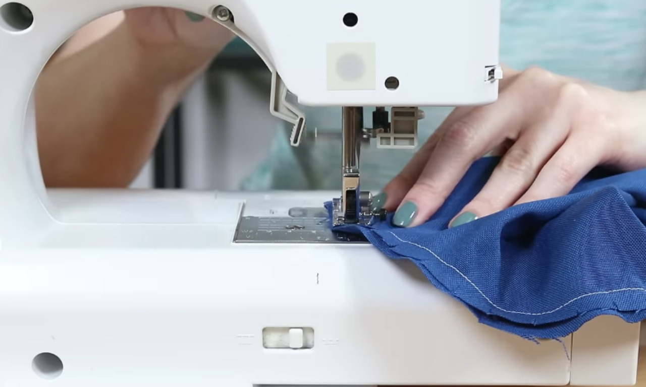 A person's hands guiding a piece of blue fabric under a sewing machine needle, with the sewing machine's presser foot holding the fabric in place. The sewing machine is white, and the person's nails are painted green.