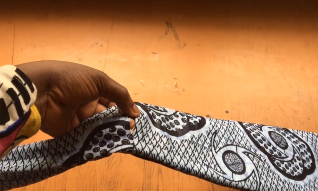 A hand holding a piece of black and white patterned fabric with intricate circular designs and geometric patterns. The fabric appears to be traditional African textile or wax print cloth. The background shows an orange-toned floor or surface. The person's wrist is partially visible, showing a colorful striped wristband or bracelet with yellow and other colors.