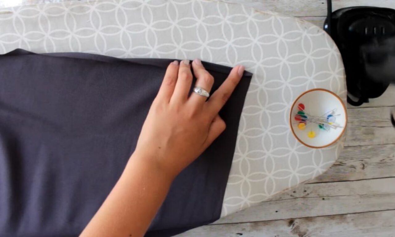 A bird's-eye view shows a hand with a silver ring carefully folding over the edge of a piece of dark, smooth fabric laid flat on a grey ironing board with a white geometric pattern. To the right of the fabric, a small white bowl with an orange rim holds several colorful headed pins. Further right, the top part of a black iron is visible resting on the end of the ironing board.