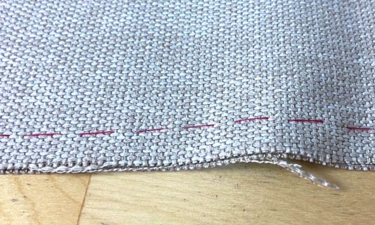 a piece of gray fabric with a textured weave, featuring three thin, parallel pink stitching lines running horizontally across the material. The fabric is laid on a light wooden surface.