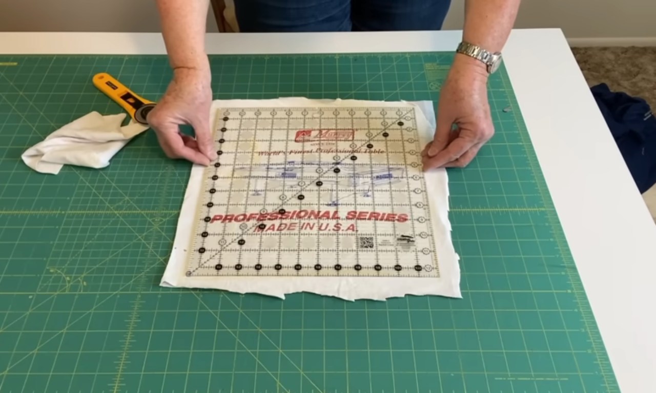 A person is using a rotary cutter to trim the edges of a piece of fabric placed on a green cutting mat. The fabric appears to be a stabilizer sheet with a grid pattern and text that reads 'PROFESSIONAL SERIES MADE IN U.S.A.' A yellow-handled rotary cutter is positioned on the mat, and a white cloth is also visible on the mat. The person is wearing a watch on their left wrist.