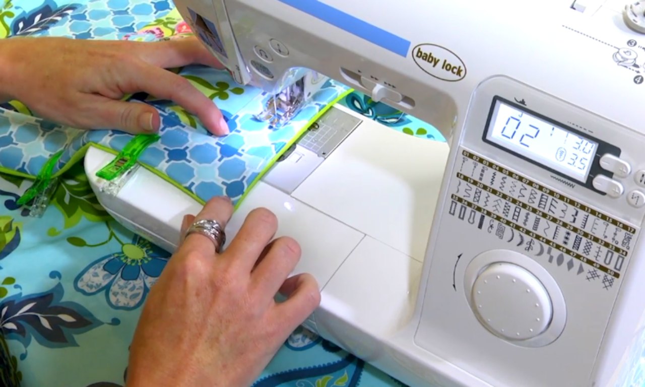 a person's hands sewing a blue and green patterned fabric on a white Baby Lock sewing machine. Green clips are holding the fabric in place as it passes under the needle.