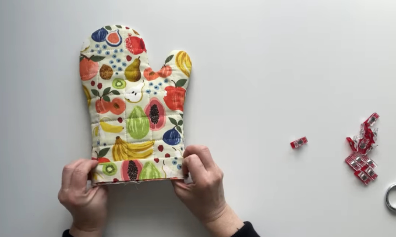 a pair of hands holding up a colorful, quilted oven mitt. The oven mitt is decorated with a playful pattern featuring various fruits such as apples, pears, bananas, kiwis, and figs. The background is a plain, light-colored surface, and there are a few scattered items nearby, including what appears to be small red and white candy wrappers and a metal ring. The overall scene suggests a crafting or sewing project.