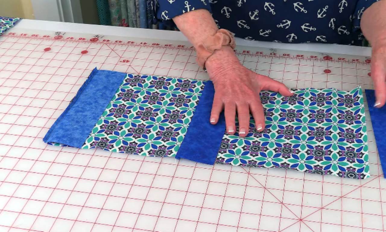 A person's hands are arranging two pieces of fabric on a cutting mat with a grid pattern. The fabric pieces have a patterned design with green, blue, and purple elements. The person is aligning the edges of the fabric pieces, likely preparing to cut or sew them together. The cutting mat has measurement markings along the edges.