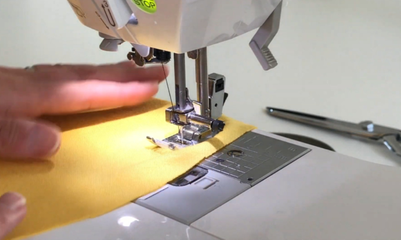 a sewing machine stitching a piece of yellow fabric, with a hand guiding the material under the needle. A pair of metal scissors lies in the background on a white surface.