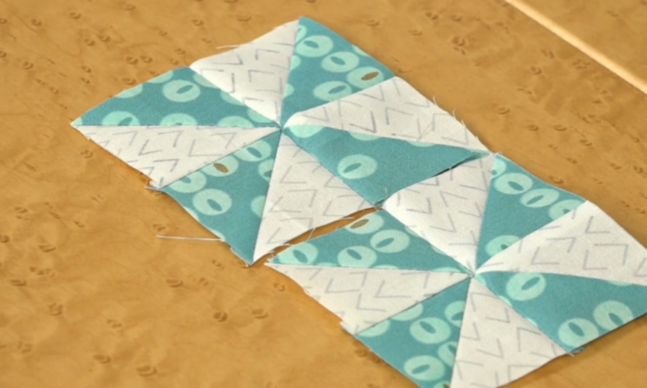 Three square quilt blocks featuring a pinwheel design lie on a light wood grain surface. Each block consists of eight triangles, alternating between a teal fabric with light blue oval patterns and a white fabric with thin grey angular lines. The blocks are sewn together horizontally, forming a row.