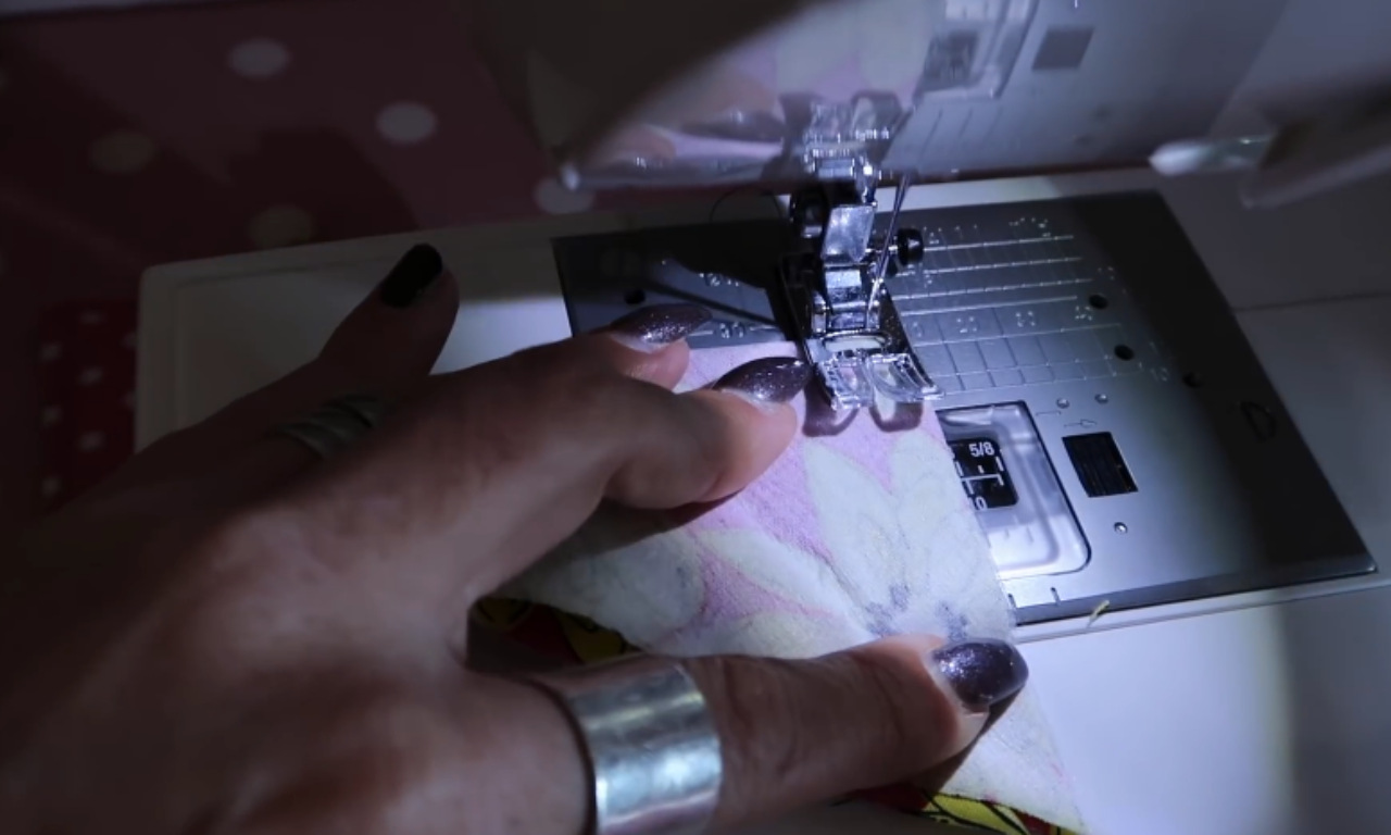 a person's hand guiding a piece of patterned fabric through a sewing machine. The person's fingernails are painted dark, and they are wearing a silver ring. The sewing machine needle is in the process of stitching the fabric, and the work area is well-lit.