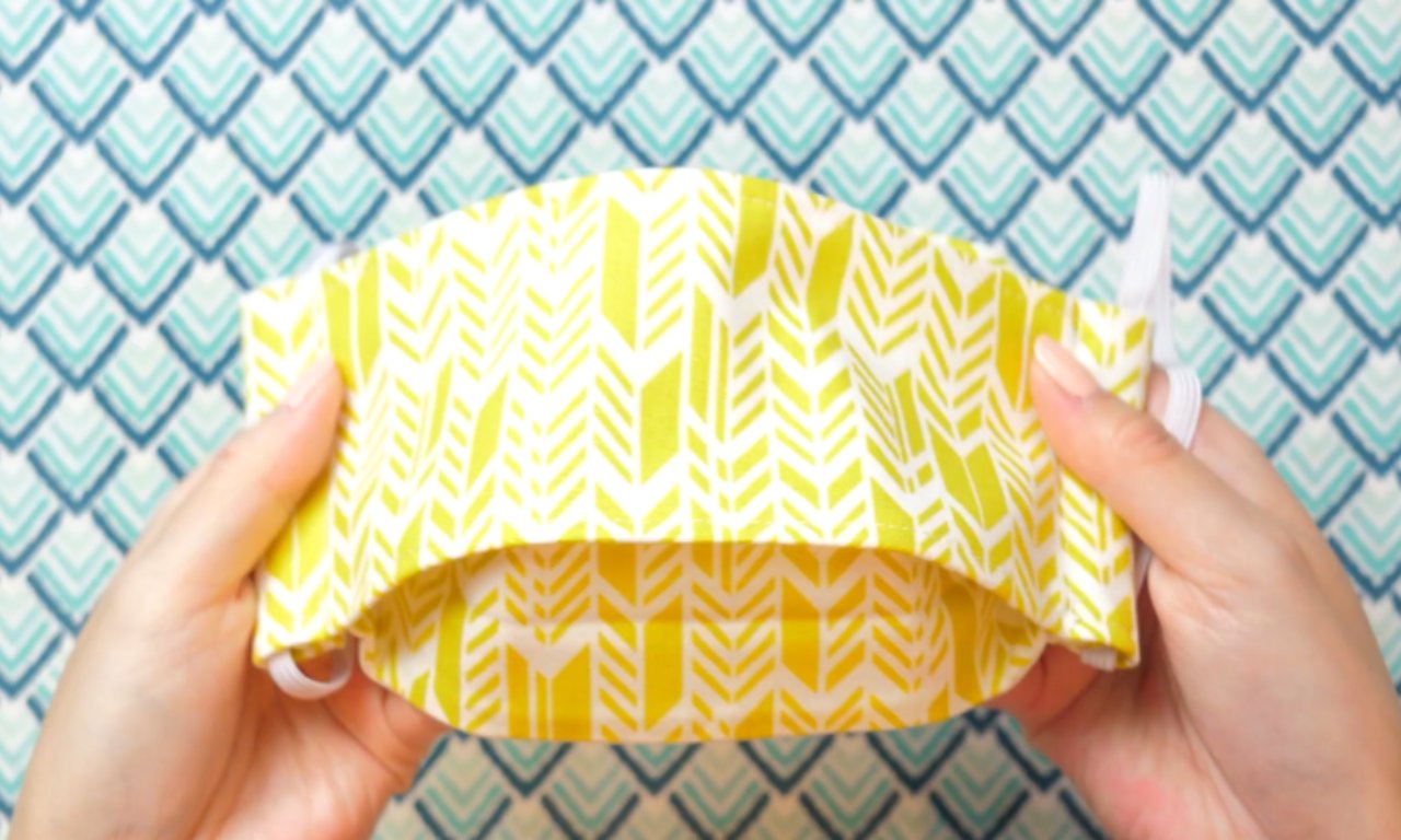 Two hands hold up a pleated yellow fabric face mask with a white arrow pattern and white elastic ear loops. The background is a blue and white scale pattern.