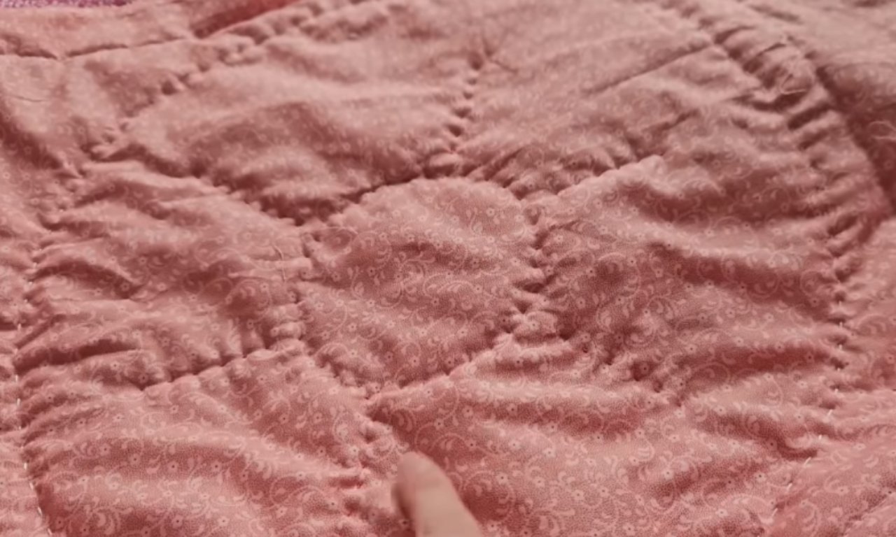 a dusty rose quilt with a subtle white paisley pattern, stitched in a grid design. The fabric appears slightly rumpled, with a few creases visible. A finger is partially visible at the bottom, touching the quilt. The overall effect is cozy and inviting.