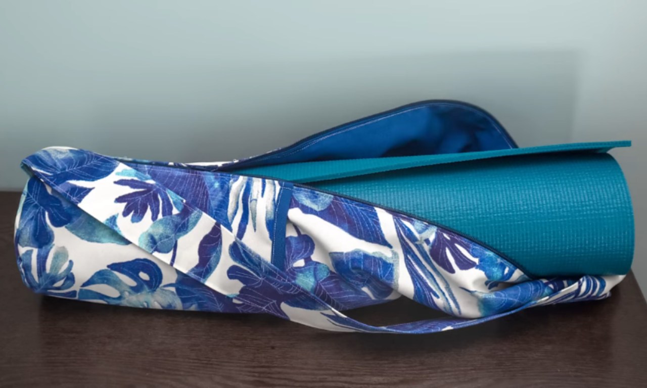 A blue and white tropical print yoga mat bag holding a rolled up teal yoga mat sits on a dark brown wooden surface against a light blue wall.